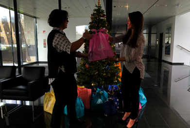 Dve ženski si pred božičnim drevescem, okrog katerega so darila, podajata HOFER vrečko.