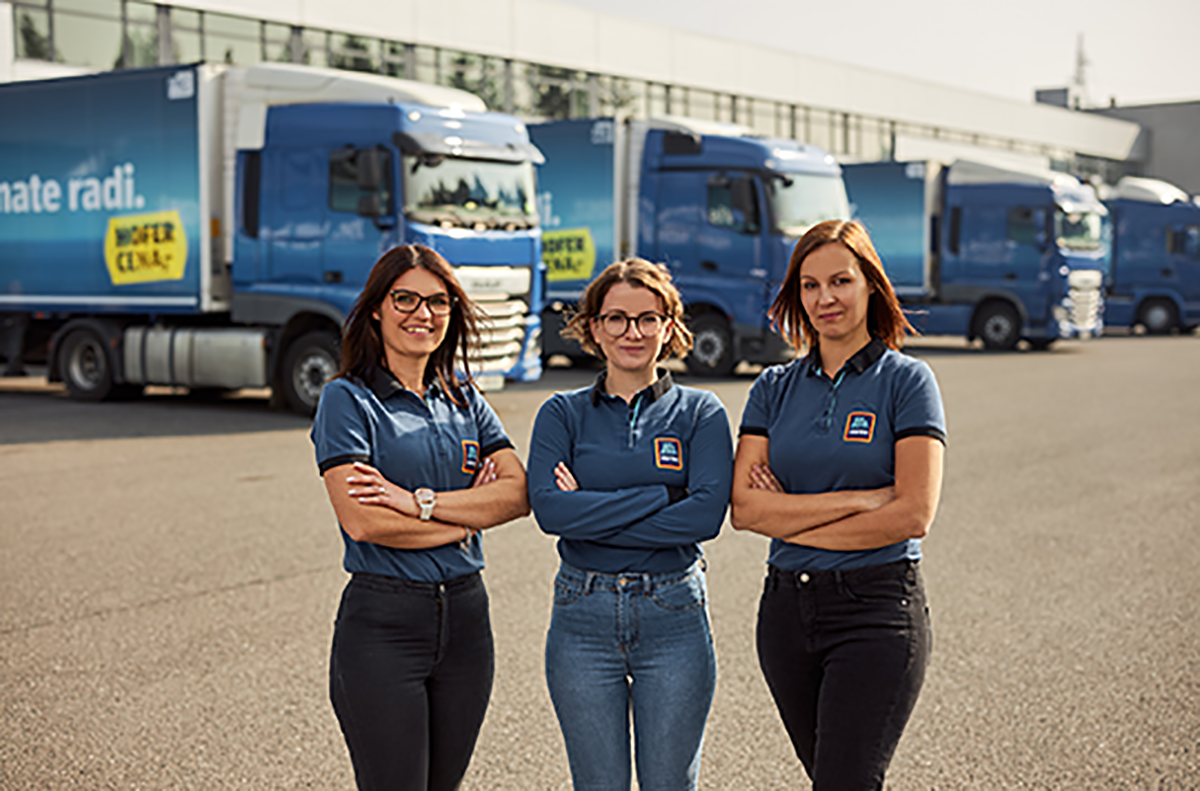 Tri sodelavke stojijo pred tovornjaki v logističnem centru in imajo prekrižane roke.