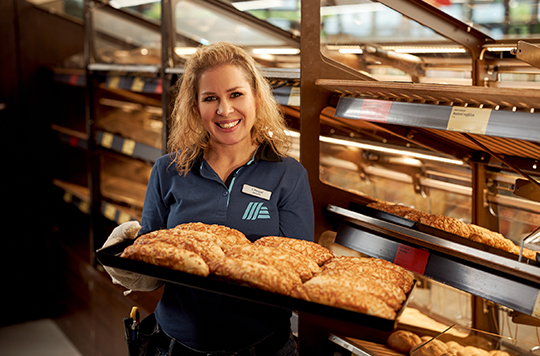 Prodajalka v pekarni drži v rokah pladenj s sirovimi žemljicami.