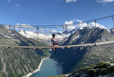 Sodelavka sedi na visečem mostu, obkrožena z gorami.